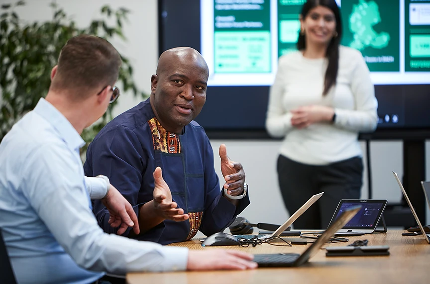 Three people can be seen – two are talking in front of a laptop, whilst a third person is standing in the background in front of a screen.
