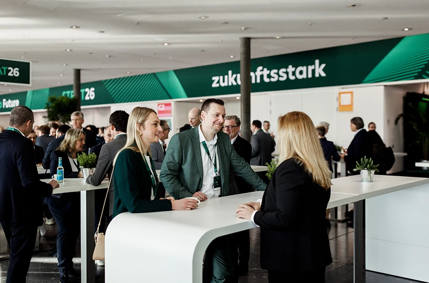 Two women and a man are standing together at a table; they look attentive and are presumably at a trade fair.