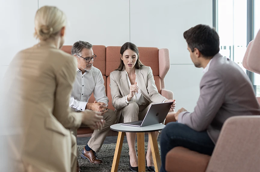 Zwei Männer und zwei Frauen sitzen im Kreis, während zwei von ihnen gemeinsam auf einen Laptop schauen.